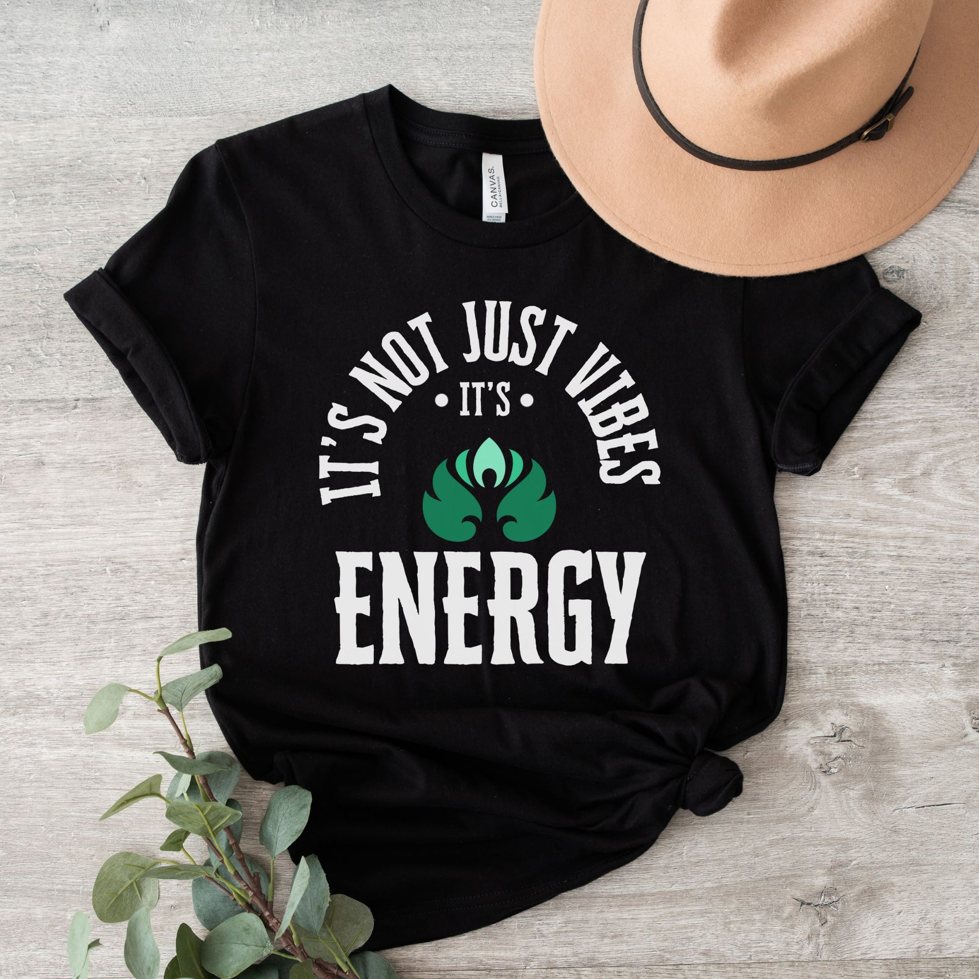 Black t-shirt with 'It's not just vibes, it's energy' text on a wooden surface with a hat and plant – spiritual and cosmic apparel by Lunar Lightwear