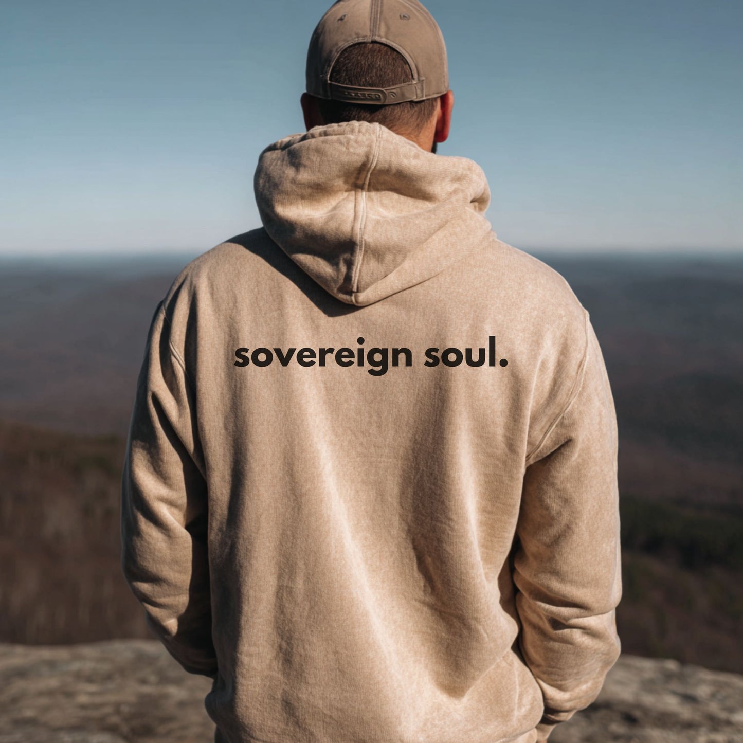 Person wearing a hoodie with 'sovereign soul.' text, standing on a mountain overlooking a scenic landscape.